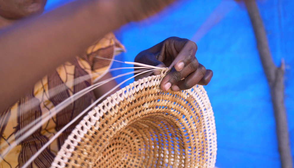 Hands weaving a circular basket from natural fibers against a blue background